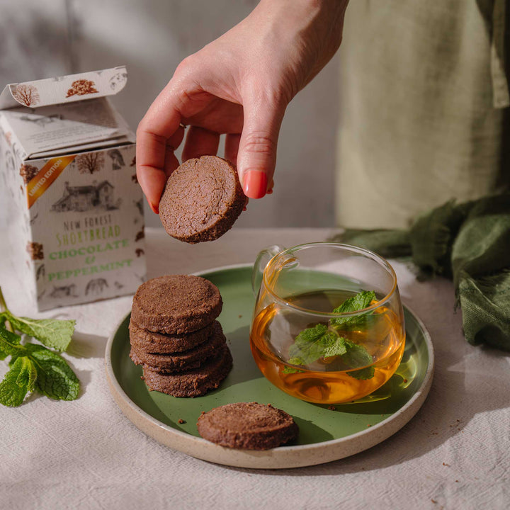 Hand holding a chocolate and peppermint shortbread cookie over a plate with shortbread and a cup of tea, next to a box of New Forest Shortbread Chocolate and Peppermint.