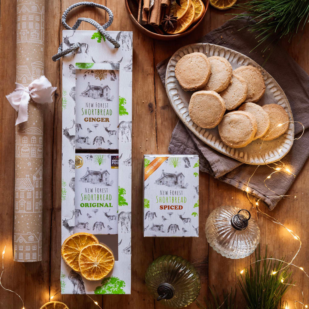 A styled flat-lay of New Forest Shortbread and their packaging on a wooden table, surrounded by Christmas decorations including fairy lights, dried orange slices, pine branches, and a rolled beige gift wrap.