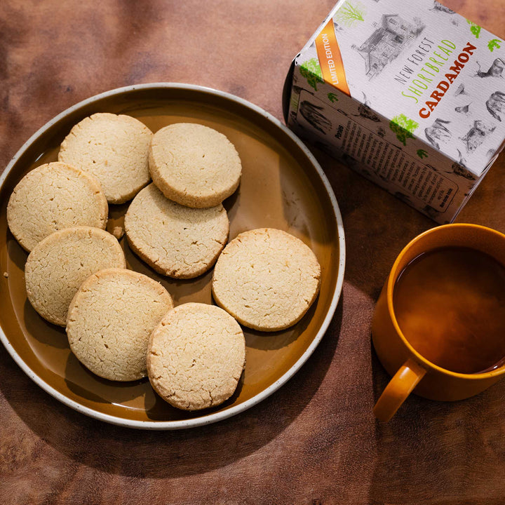 Cardamon shortbread cookies on a plate with a mug of coffee and a box of shortbread in the background on a dark brown surface.