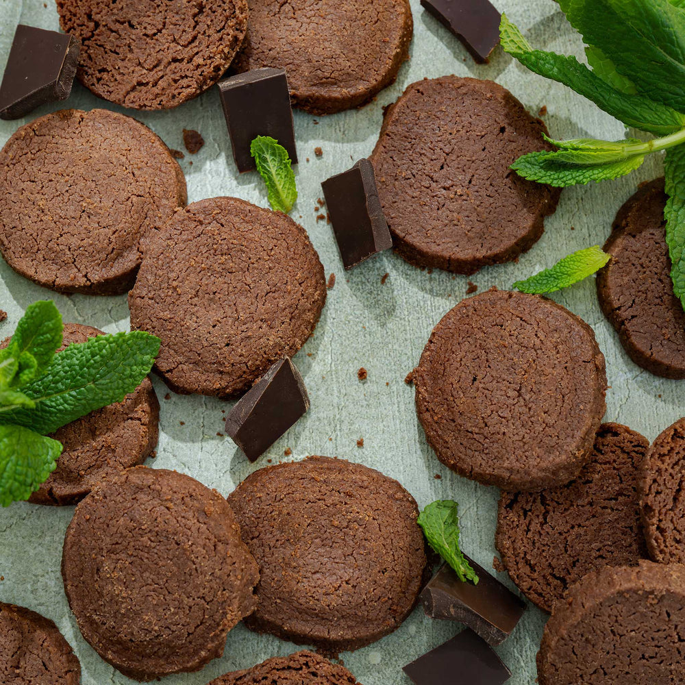 Chocolate shortbread cookies with mint leaves and chunks of chocolate on a textured surface