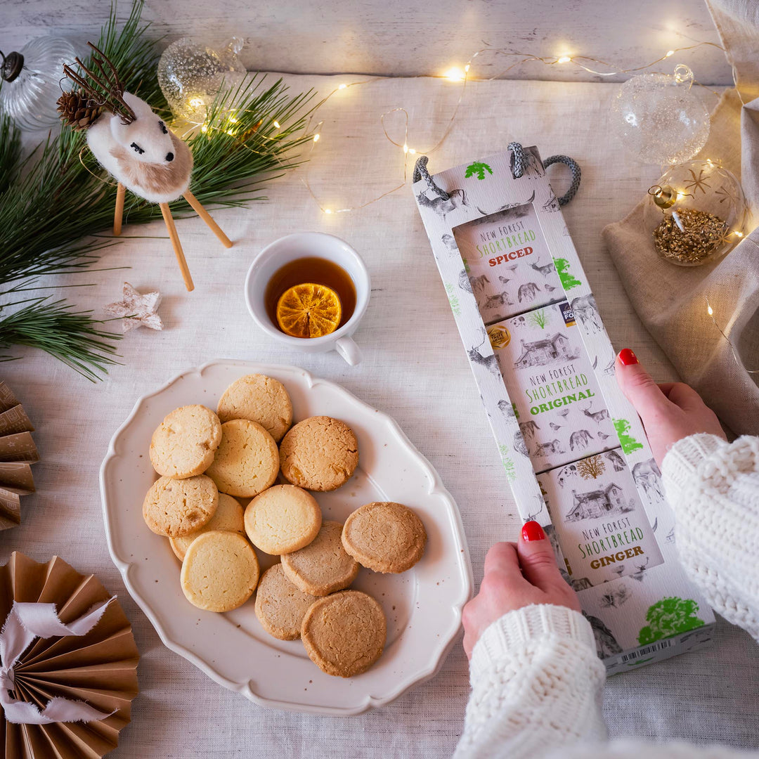 A styled Christmas flat-lay featuring three boxes of Christmas Shortbread being held by hands with red nail polish, next to a plate of shortbread cookies and a cup of tea with a lemon slice, decorated with fairy lights and pine branches.