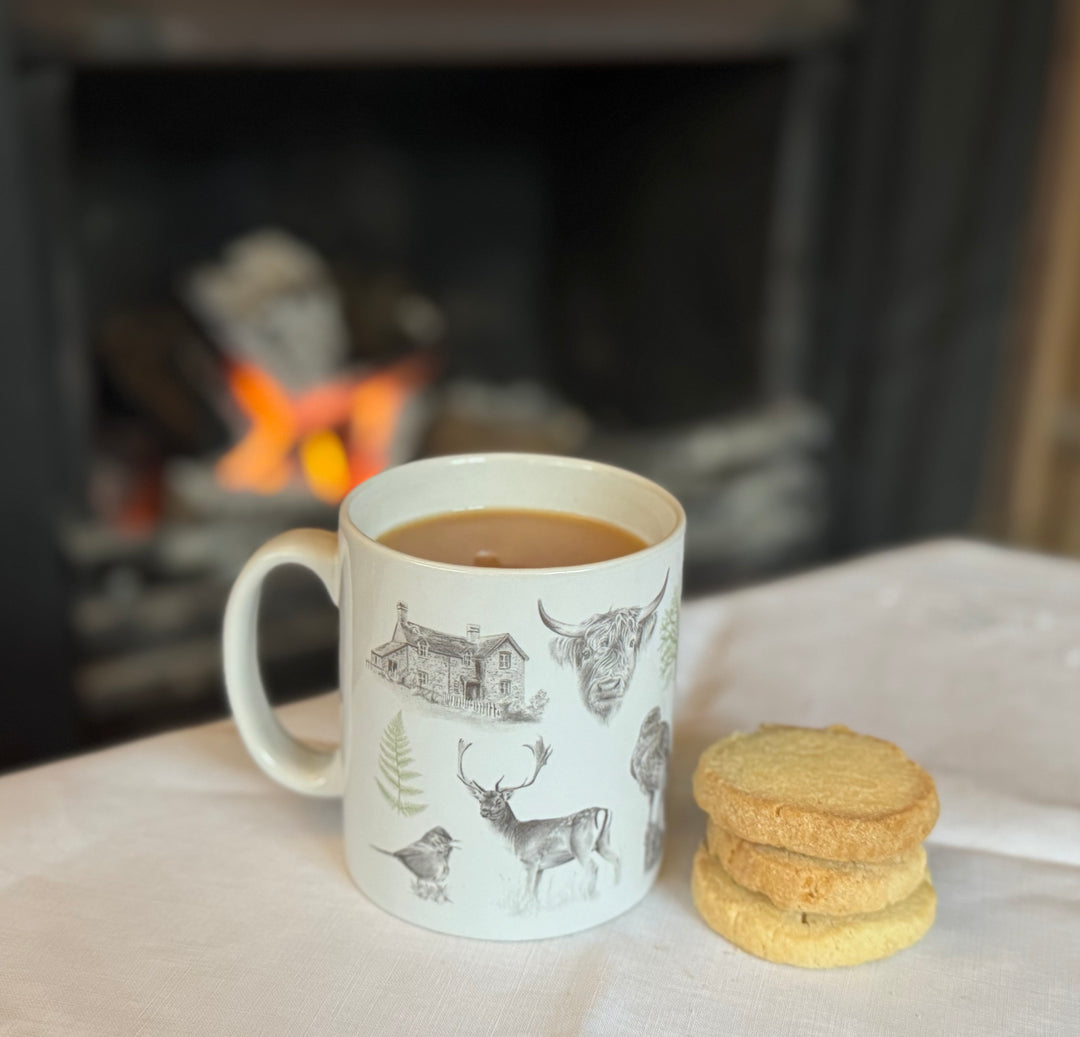 A white ceramic mug with illustrations of animals and Burley Rails cottage, placed on a table with a stack of shortbread cookies in front and a fire in the background.