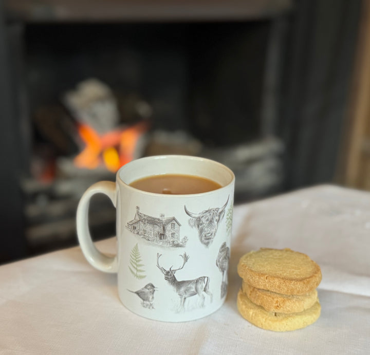 A white ceramic mug with illustrations of animals and Burley Rails cottage, placed on a table with a stack of shortbread cookies in front and a fire in the background.