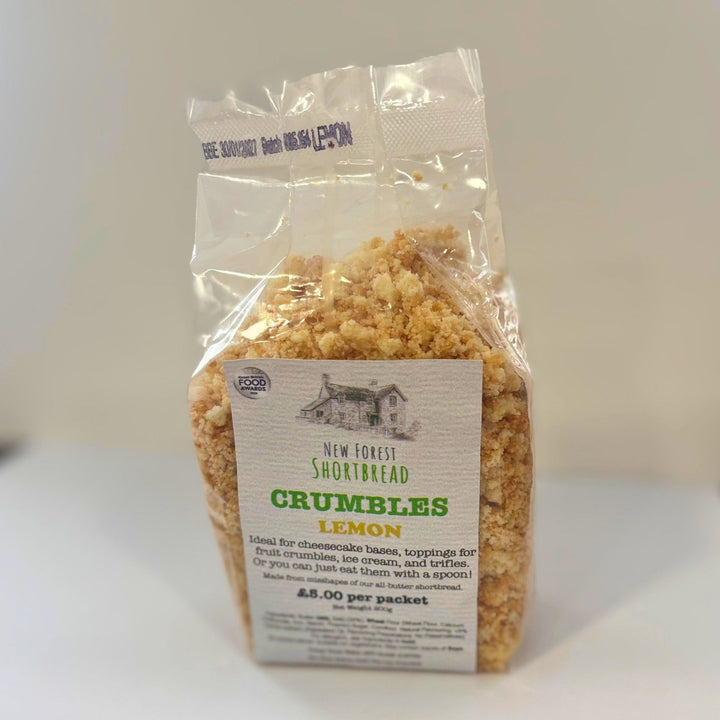 A clear plastic bag of New Forest Shortbread Crumbles Lemon, showing the golden-brown crumb contents and a white paper label with green and black text, set against a plain light gray background.