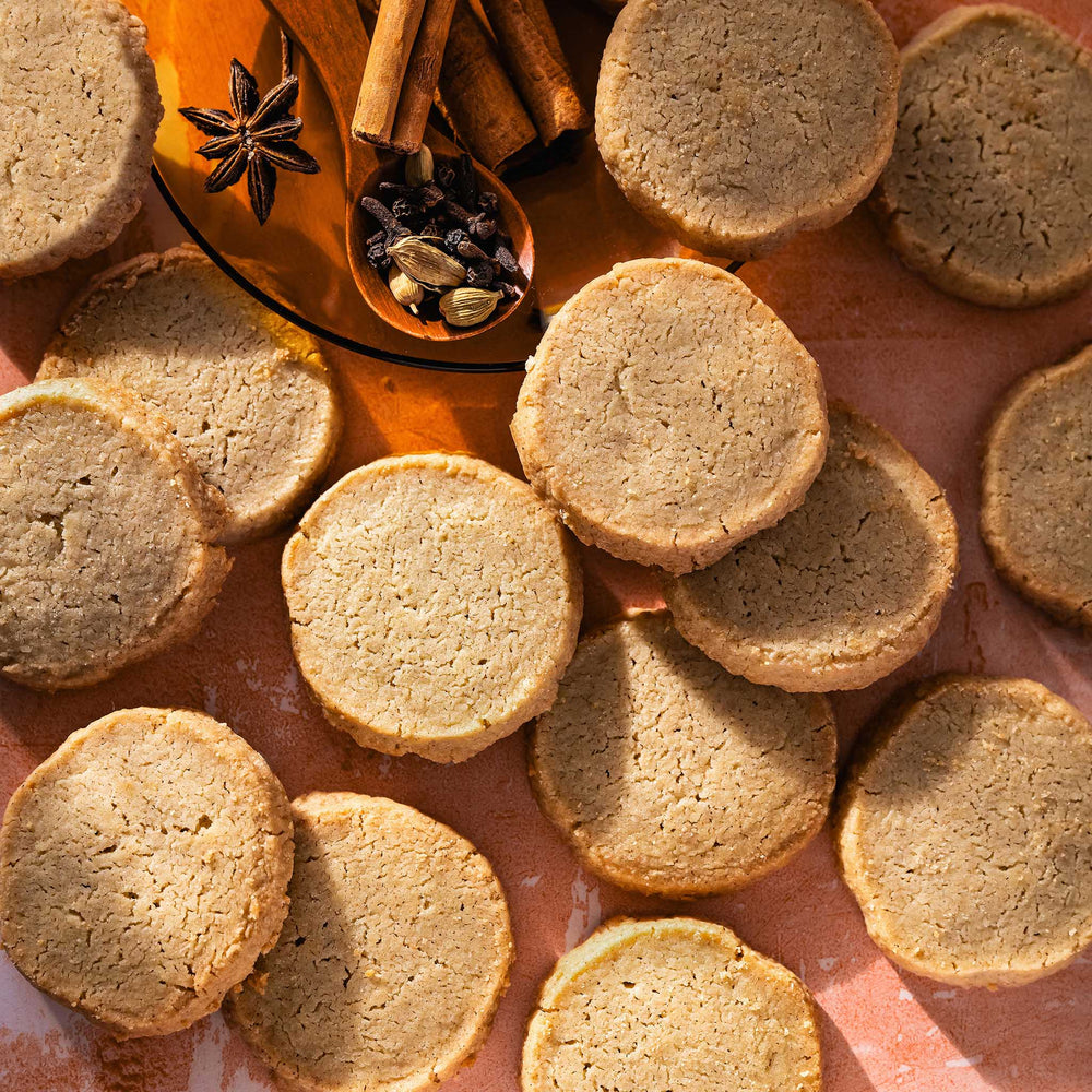Round golden-brown spiced shortbread arranged on a warm pinkish surface, with cinnamon sticks, star anise, and spices in a glass dish in the background.