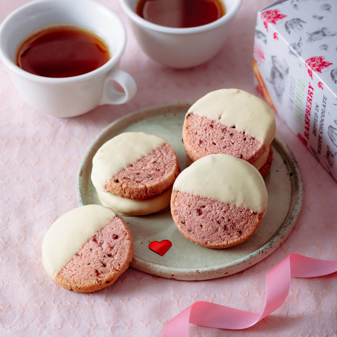 Pink and white shortbreadon a plate with two cups of tea and a box of raspberry and white chocolate shortbread in the background.