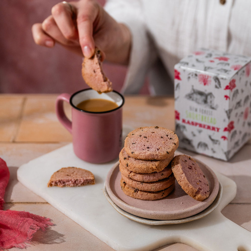 Person dipping a raspberry shortbread cookie into a cup of tea with a stack of shortbread pieces on a plate.