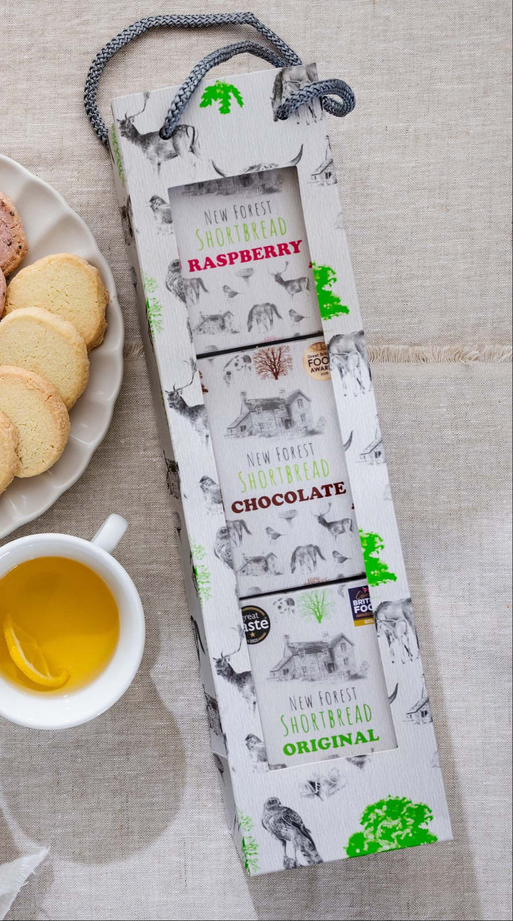 Assorted shortbread on a plate with two cups of tea and a selection bag of New Forest shortbread on a light surface.