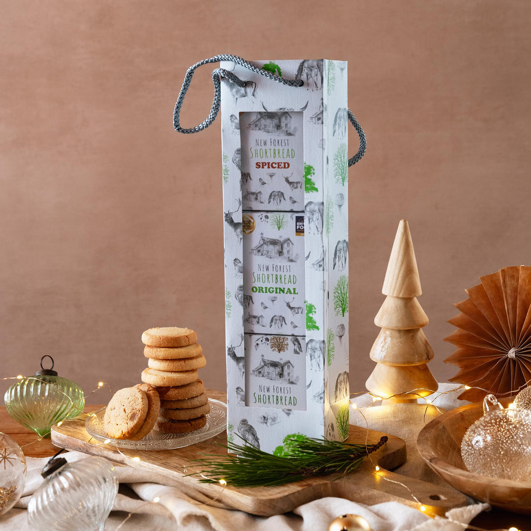 A tall white gift bag of Original, Ginger and Spiced Shortbread Flavours stands on a wooden board in a styled festive setting with shortbread cookies, string lights, and decorative ornaments against a warm beige background.